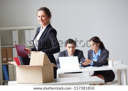 Portrait of smart businesswoman with box looking at camera on background of working colleagues