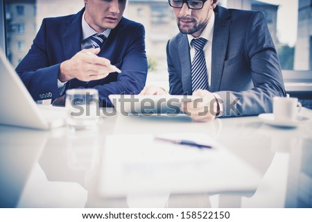 Image of two young businessmen discussing document in touchpad at meeting