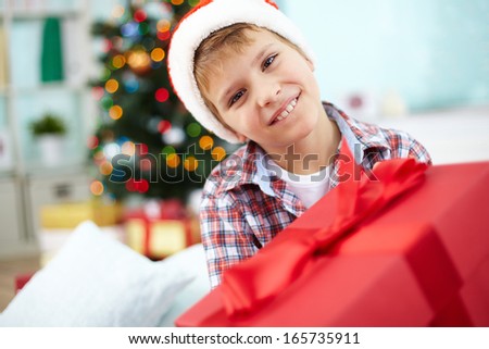 Portrait of cheerful boy with big red giftbox looking at camera on Christmas evening