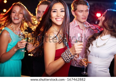 Portrait of cheerful girl with champagne flute dancing at party while smiling at camera