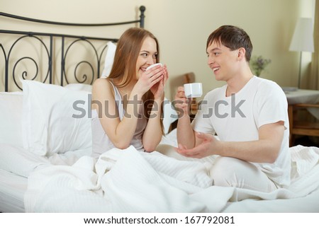 Happy young couple sitting in bed and looking at one another while drinking tea