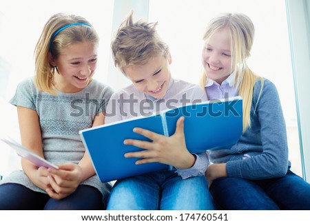 Portrait of three laughing classmates looking at notes in exercise book of schoolboy while he writing