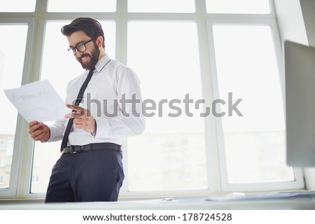 Smiling businessman standing by the window in office and reading paper