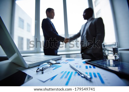 Image of eyeglasses, glass of water, pen, laptop, touchpad and financial documents at workplace with businessmen handshaking on background