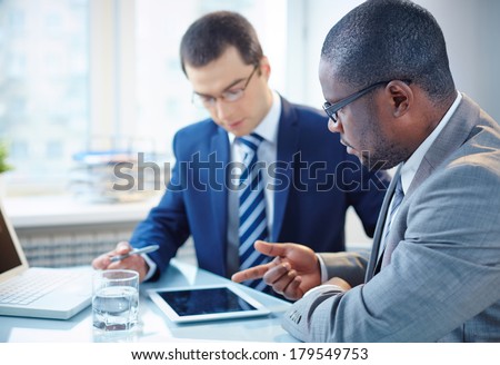 Image of two young businessmen discussing data in touchpad at meeting