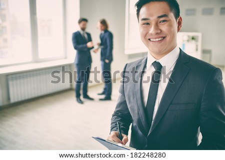 Close-up portrait of a young successful manager with clipboard posing at camera while his colleagues talking on the background