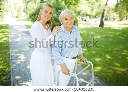 Pretty nurse and senior patient with walking frame looking at camera outside