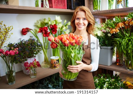 Portrait of young female florist with big vase of red tulips looking at camera