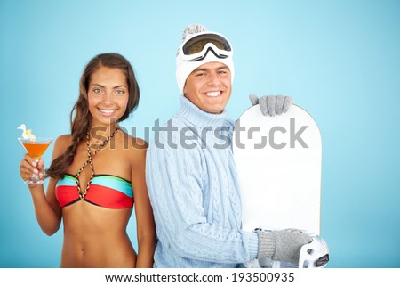 Portrait of cute girl in bikini holding cocktail and handsome man in pullover holding snowboard