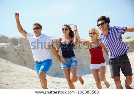 Ecstatic young people holding by hands while running down sandy beach
