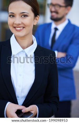 Image of pretty businesswoman with cellphone looking at camera on background of her co-worker