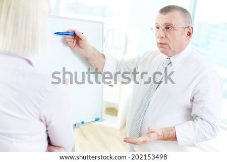 Image of mature businessman teaching his partner on whiteboard at meeting