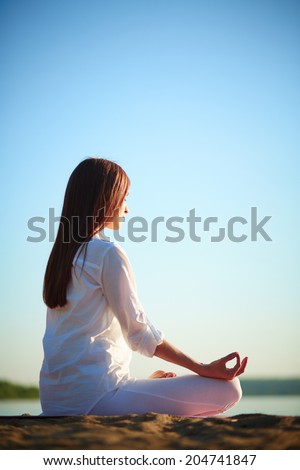 Side view of meditating woman sitting in pose of lotus against blue sky outdoors