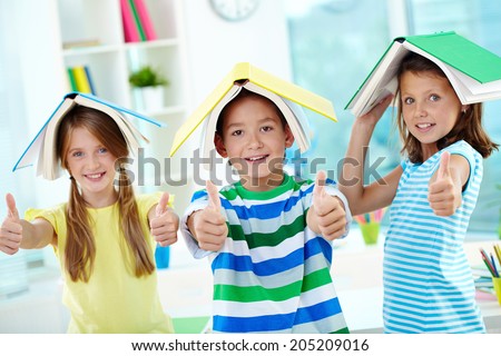 Portrait of happy classmates with books on their heads keeping thumbs up