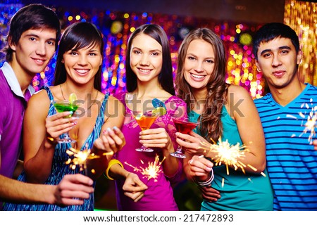 Company of happy girls and guys looking at camera while toasting with cocktails at New Year party