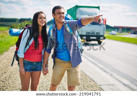 Couple of young hitch-hikers standing by highway while guy pointing somewhere