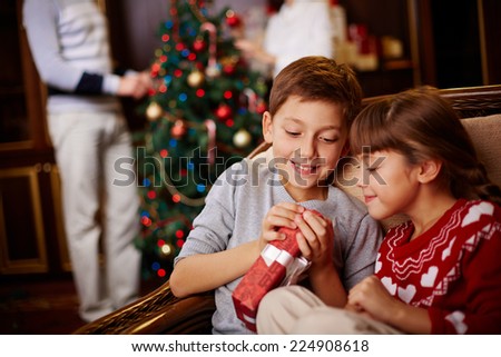 Two siblings opening Christmas gift with their parents decorating fir-tree on background