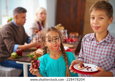 Portrait of smudgy siblings eating sweet pie and looking at camera with their parents on background