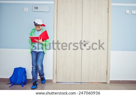 Cute schoolboy writing notes in exercise-book while standing by classroom door