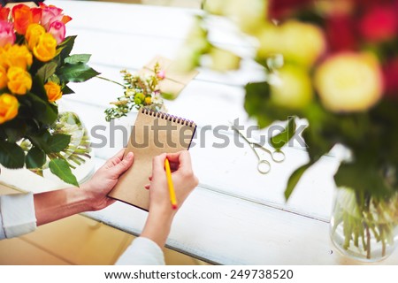 Hands of florist with pencil writing in notepad on work day