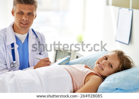 Little girl lying in bed and looking at camera with her doctor on background