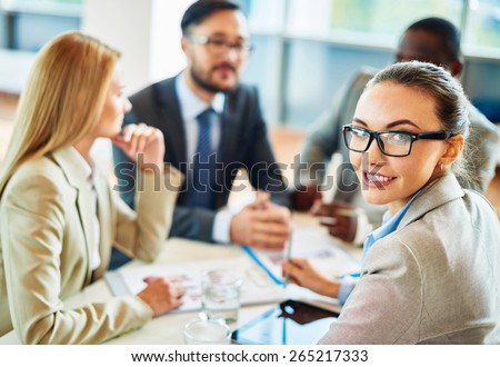 Smiling secretary in eyeglasses looking at camera in working environment