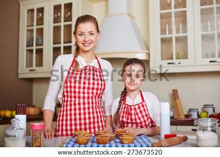 Pretty girl and her mother looking at camera with baked muffins on table near by