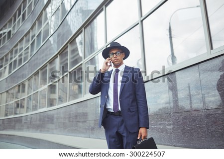 Young man in suit, hat and eyeglasses speaking on the phone while standing by modern building