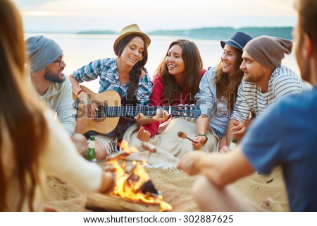 Friendly girls and guys singing by guitar by campfire