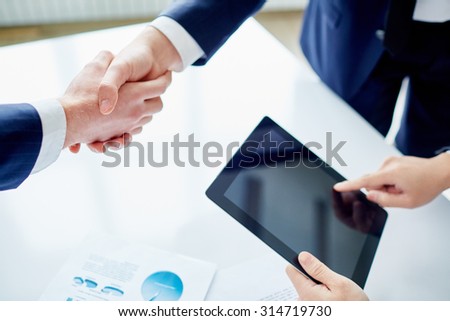 Partners shaking hands while businessman with touchpad networking near by
