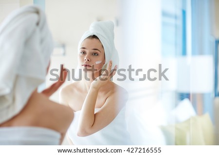 Young woman applying foundation or moisturizer on her face in front of mirror