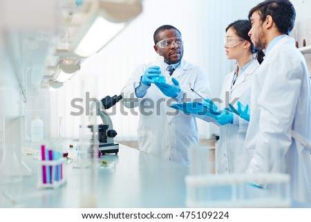 Male African-American laboratory scientist holding flask with blue liquid discussing chemical reaction with male Latin-American and female Asian colleagues.