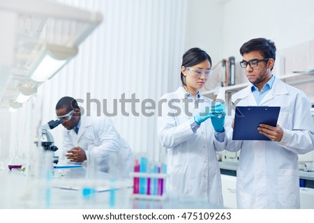 Concentrated Asian female laboratory scientist in safety goggles holding flask with blue liquid showing it to Latin-American colleague writing down results. African-American scientist in background.
