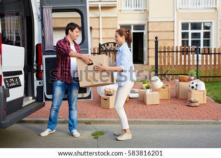 Portrait of man taking cardboard boxes out of moving van and passing them to his wife in front of new house