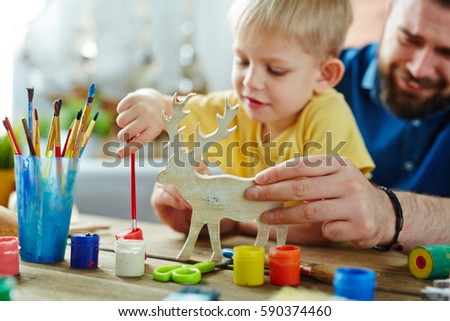 Little blond-haired son with his bearded dad preparing Christmas present for granny: they sitting at table and enthusiastically coloring toy deer in different colors