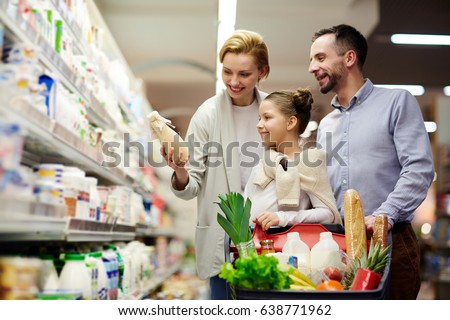 Portrait of happy family shopping in grocery store: choosing milk in dairy product department with cart full of food