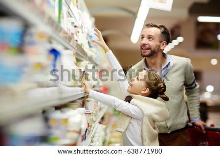 Happy family grocery shopping in supermarket: smiling man with daughter choosing dairy products from fridge in milk aisle