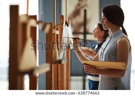 Side view portrait of young female student in art class, enjoying painting oil picture on canvas