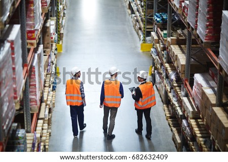 Group of warehouse workers wearing hardhats and reflective jackets waking in aisle between tall racks with packed goods, back view