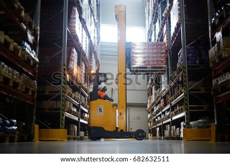 Side view  portrait of warehouse worker using reach fork truck to load pallet with boxes on tall rack