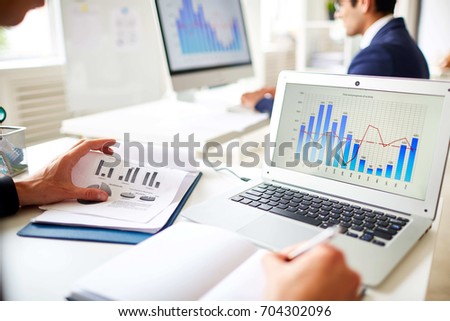 Open plan office wrapped up in work: unrecognizable manager taking notes while analyzing statistic data while his male colleague preparing financial accounts on modern computer