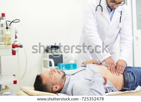 Concentrated young physician doing abdominal palpation of middle-aged patient suffering from pain, interior of modern office on background