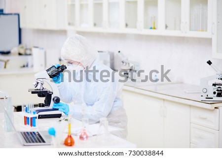Profile view of female microbiologist wearing hazmat suit looking through microscope while studying mutation virus at modern laboratory