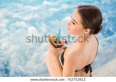 Slim girl with tropical cocktail sitting by swimming-pool with delicate waves of warm water