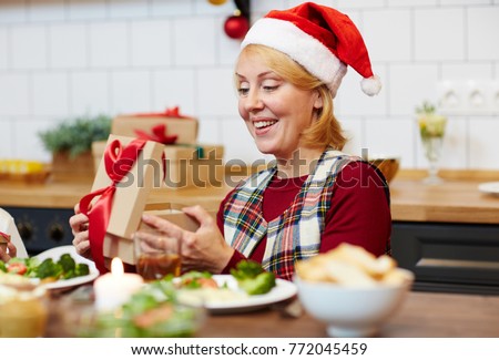 Smiling female sitting by festive table and looking at surprise in gift-box during xmas dinner