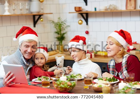 Family in Santa caps gathered by festive table to have dinner and watch online xmas video