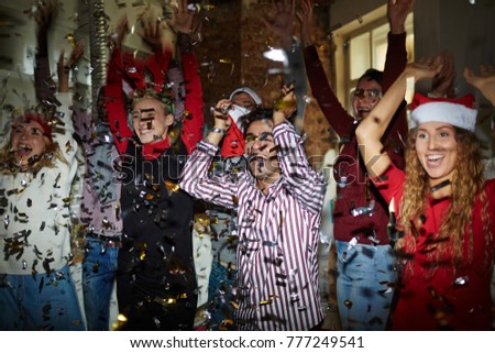 Excited young friendly people with raised hands dancing at xmas party in confetti fall