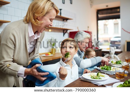 Little boy eating steamed broccoli and looking at his grandmother during dinner in the kitchen