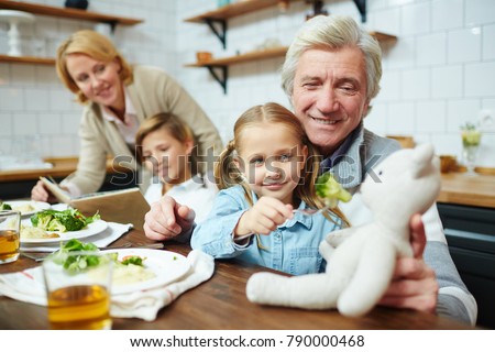 Happy mature man holding teddybear while his granddaughter giving food to her toy by dinner