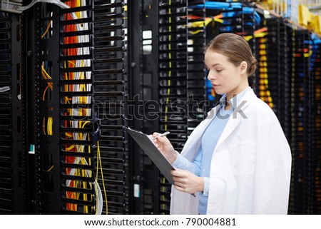Female technician or expert in whitecoat making working notes in data center